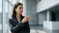 Professional Asian businesswoman in dark suit gesturing while presenting in modern office environment with natural lighting and contemporary architecture.
