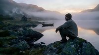 Man sitting on rocky shore contemplating misty mountain lake at dawn with wooden dock and serene atmosphere for meditation and outdoor adventure concepts.