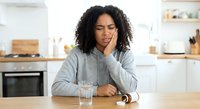 Young African American woman experiencing tooth pain holding cheek in modern kitchen with glass of water on wooden table for dental health concept.
