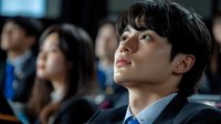 Young Asian businessman in suit attentively listening during corporate conference or seminar with blurred audience in background.