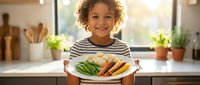 Happy mixed-race child holding healthy vegetable plate in modern kitchen. Young boy with curly hair presenting colorful meal for nutrition education.