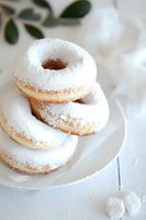 Fresh powdered sugar donuts stacked on white plate against light background, soft focus with delicate bokeh effect and green plant detail. Food photography.