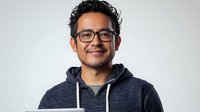 Young Hispanic male professional wearing glasses and casual hoodie smiling confidently against light background. Portrait conveys warmth, approachability and modern workplace culture.