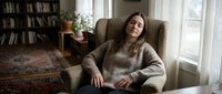 Woman with long brown hair and fair complexion, wearing a cozy knit sweater, sitting relaxed in an armchair in a bright, modern living room with bookshelves and natural light.