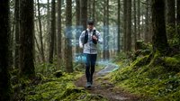 Young Caucasian male hiker walking on forest trail with backpack among tall trees and moss-covered ground for outdoor adventure and nature exploration.