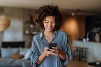 Young African American woman with curly hair smiling while using smartphone in cozy modern apartment with warm lighting. Perfect for lifestyle and technology content.
