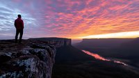 Male hiker silhouette standing on rocky cliff edge overlooking canyon river at dramatic sunrise with vibrant pink orange clouds in sky for outdoor adventure concepts.