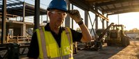 Senior construction worker in safety vest and hard hat at building site during golden hour sunset with heavy machinery and structural framework in background.
