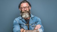 Smiling bearded middle-aged Caucasian man in denim jacket and glasses with crossed arms on gray background for business portraits and lifestyle content.