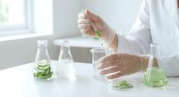 Laboratory scientist in white coat and gloves conducting plant research with green leaf samples in glass beakers and test tubes for botanical analysis.