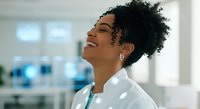 Young woman of African descent with curly black hair wearing white shirt and lanyard in modern office environment, smiling with head tilted back, expressing joy and confidence at work.