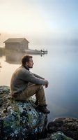 Contemplative young Caucasian man in wool sweater sitting on rocky shore at misty lake during golden hour sunrise with wooden boathouse in background.