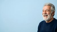 Senior man with gray hair and beard wearing navy sweater smiling against light blue background, suitable for healthcare, wellness, lifestyle and corporate portraits.