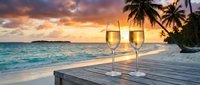 Two champagne glasses on wooden table at tropical beach during golden sunset with palm trees and turquoise ocean creating romantic paradise atmosphere.