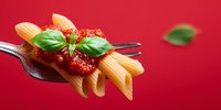 Penne pasta with tomato sauce and fresh basil on fork against vibrant red background, classic Italian cuisine presentation.