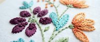 Colorful hand embroidered floral design on white fabric featuring purple, teal and orange flowers with green stems in traditional needlework style.