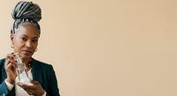 Confident African American woman with braided updo hairstyle wearing dark blazer and earrings, holding business card against neutral beige background for corporate and professional use.