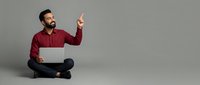Man in maroon shirt sitting cross-legged with laptop, pointing upward against gray background. Suitable for business, technology, education, and online learning concepts.