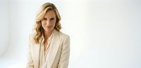 Professional blonde businesswoman in elegant beige suit jacket smiling confidently against white background for corporate marketing materials.