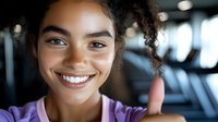 Young African American woman with curly hair and bright smile wearing purple athletic top at fitness center, closeup portrait with blurred gym background.