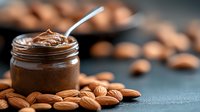 Creamy chocolate almond butter in glass jar with spoon and scattered raw almonds on dark background, macro food photography for healthy eating blogs.