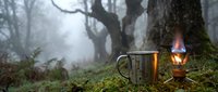 Steaming metal mug and vintage lantern create cozy camping atmosphere in misty forest with moss-covered ground and bare trees for outdoor adventure content.