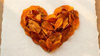 Heart-shaped arrangement of dried autumn leaves in warm orange and brown tones on textured paper background for seasonal crafts and romantic designs.