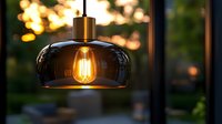Modern pendant lamp with glowing vintage filament bulb in smoked glass shade against blurred sunset garden background, creating warm atmospheric lighting.