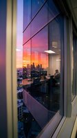 Modern office interior with floor-to-ceiling windows overlooking vibrant sunset cityscape, silhouetted businesspeople working at desks, urban skyline with illuminated buildings, golden and pink sky reflection on glass.