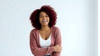 Happy young African American woman with curly red hair laughing joyfully in pink cardigan against white background for lifestyle and wellness content.