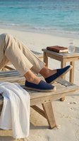 Man relaxing on sandy beach wearing navy espadrilles, linen pants, seated on wooden lounger with book and water glass by turquoise ocean.