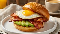 Gourmet breakfast burger with fried egg, crispy bacon and avocado slices on sesame bun served with coffee and orange juice for morning meal concept.