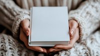 Female hands with manicured nails holding closed white hardcover book against cozy knitted sweater background, creating minimalist composition for reading and lifestyle concept.