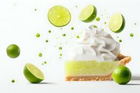 Fresh key lime pie slice with whipped cream topping, surrounded by sliced limes and lime juice drops on white background, bright food photography.