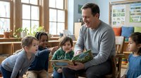 Adult male, Caucasian, teacher or educator, reading storybook to diverse group of young children in bright classroom setting with natural light from windows, colorful furniture, and educational materials on walls.