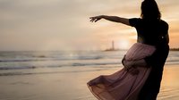 Silhouette of woman dancing gracefully on beach at sunset with flowing dress, ocean waves and golden sky creating romantic atmospheric scene.