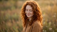 Peaceful redhead woman with curly hair smiling with closed eyes in warm autumn sunlight, expressing joy and serenity in natural outdoor setting.