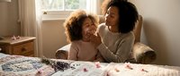 Woman with curly dark hair and young child with curly hair sitting on bed near window, engaging in bonding moment, suitable for family, parenting, and lifestyle content.