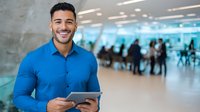 Young Hispanic businessman smiling while using digital tablet in modern office space with colleagues in background, representing corporate success.