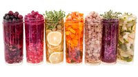 Colorful assortment of fermented vegetables and fruits in glass jars including beets, cabbage, citrus, carrots, and herbs arranged in row on white background.