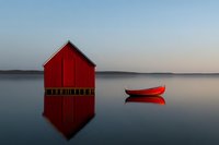 Red wooden boathouse and traditional red canoe on calm lake water at dawn, with perfect mirror reflection creating minimalist Nordic landscape scene.