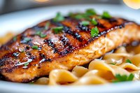 Grilled salmon fillet with caramelized glaze and char marks served on creamy pasta, garnished with fresh herbs on white plate, close-up view of gourmet meal.