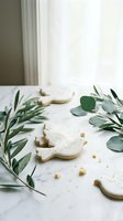 Elegant white Christmas cookies decorated with intricate icing patterns surrounded by fresh eucalyptus and olive branches on marble surface for holiday baking.