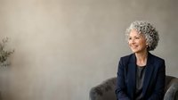 Senior woman with gray curly hair and Caucasian ethnicity wearing dark blazer, seated in modern chair against neutral background for corporate, business, and professional portrait use.