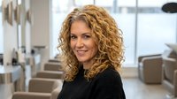 Professional blonde curly-haired woman smiling in modern office environment with natural lighting and contemporary furniture for business presentations.