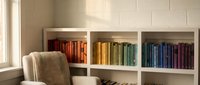 Organized home library with color-coordinated books in white shelving unit and beige armchair by window for reading and studying interior design.