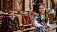 Young girl with braided hair and headpiece wearing traditional striped tunic in textile market surrounded by colorful woven rugs and carpets for sale.