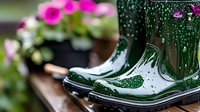 Wet green rubber boots with water drops in garden setting, pink flowers in background creating spring mood and rainy day atmosphere.