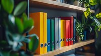Colorful office binders arranged in rainbow order on wooden shelf with green plants, creating organized workspace atmosphere for business and home office decor.