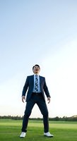 Confident Asian male businessman in navy suit and checkered tie standing with wide stance on open green field under clear sky, expressing determination and success in outdoor corporate setting.
